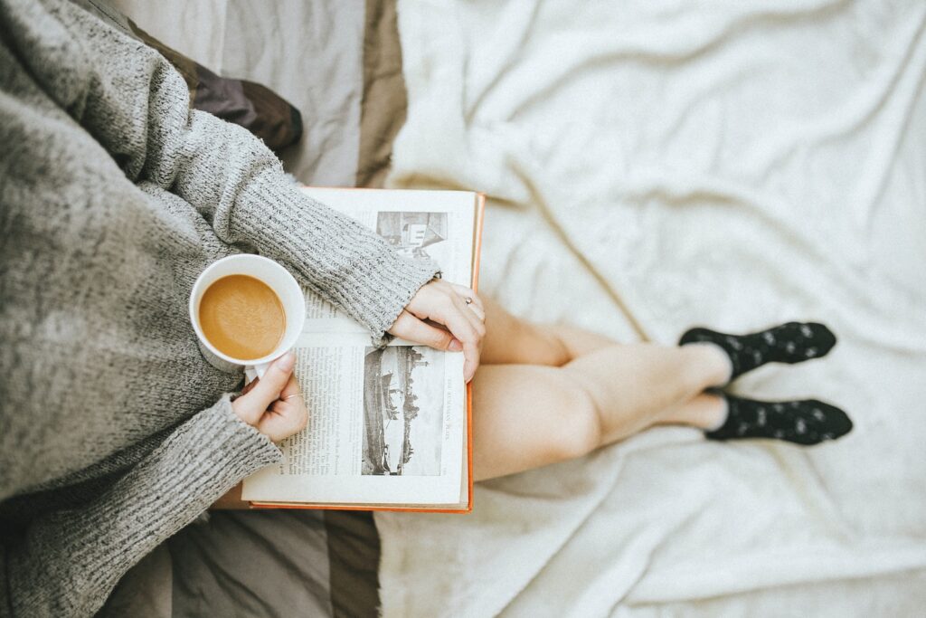 woman holding a cup of coffee at right hand and reading book on her lap while holding it open with her left hand in a well-lit room for self care kit ideas