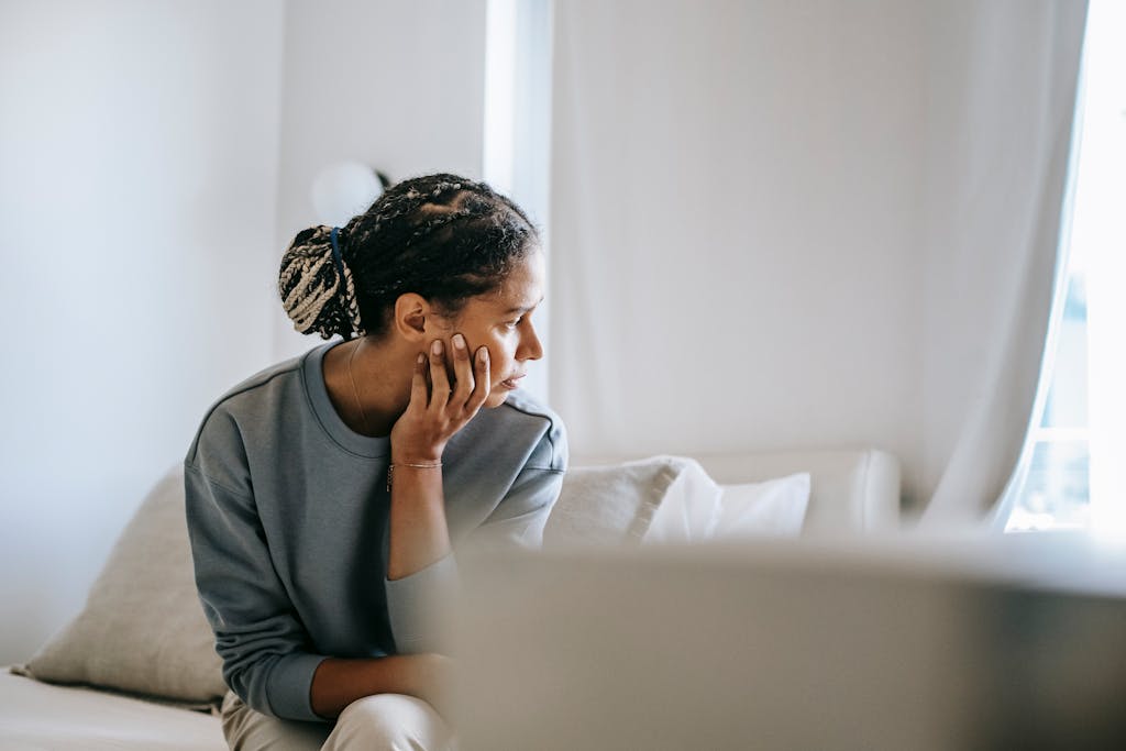 postpartum recovery for A young woman with braided hair sits pensively, gazing out the window in a tranquil, softly lit room.